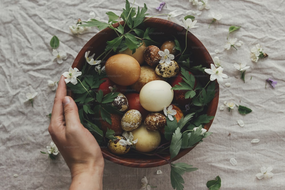 Easter eggs with spring flowers in wooden bowl and hand on rustic linen background, top view. Aesthetic seasons greeting. Stylish  easter and quail eggs in natural dye and spring blooms.