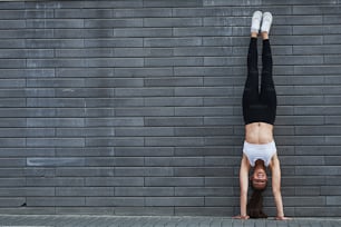 Fare la verticale. Giovane bruna sportiva con la forma del corpo sottile contro il muro di mattoni in città durante il giorno.