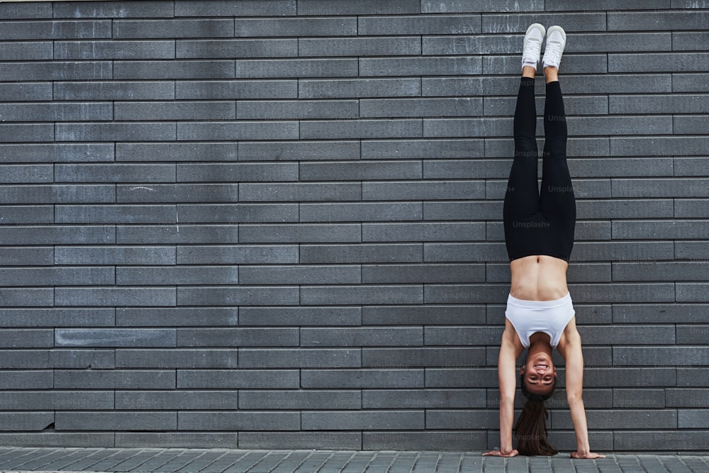 Doing handstand. Young sportive brunette with slim body shape against brick wall in the city at daytime.