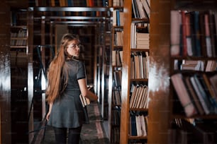 Looks behind. Female student is in library that full of books. Conception of education.