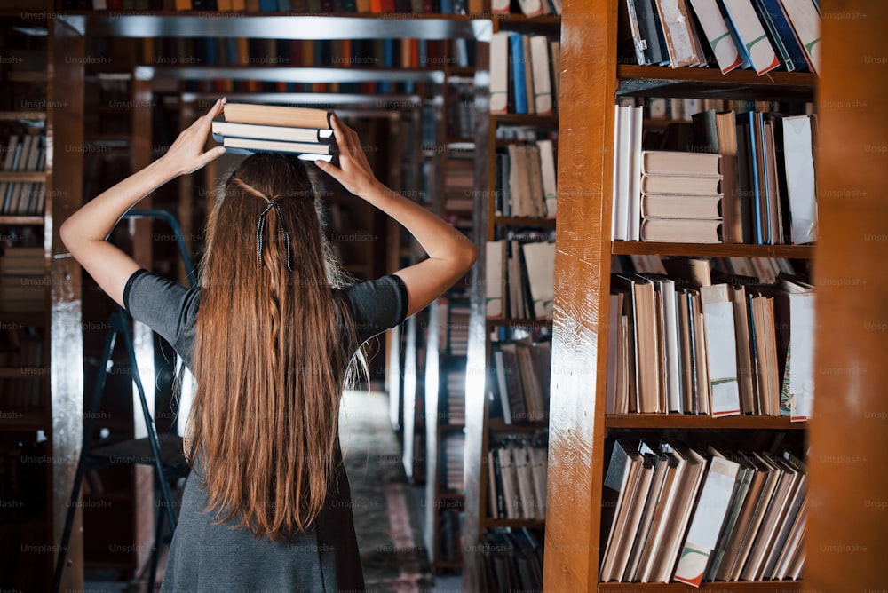 Fröhliches Mädchen hat Spaß, während sie Bücher auf ihren Kopf legt. Studentin ist in der Bibliothek. Konzeption von Bildung.