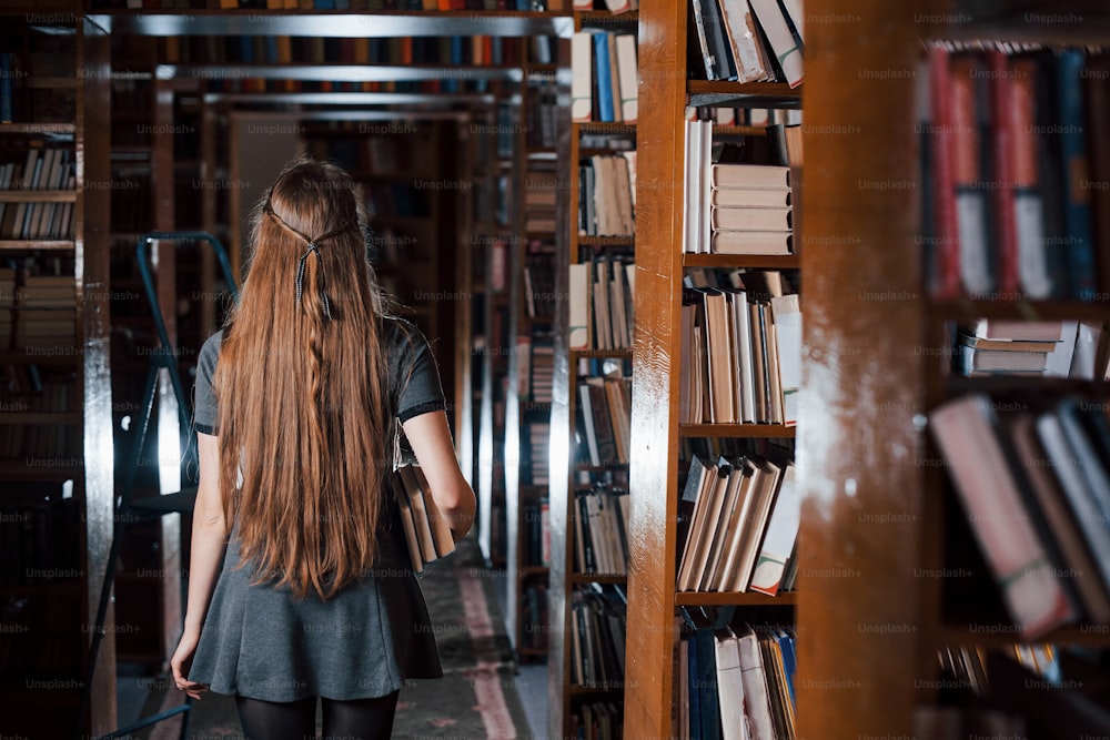 Back view. Female student is in library that full of books. Conception of education.