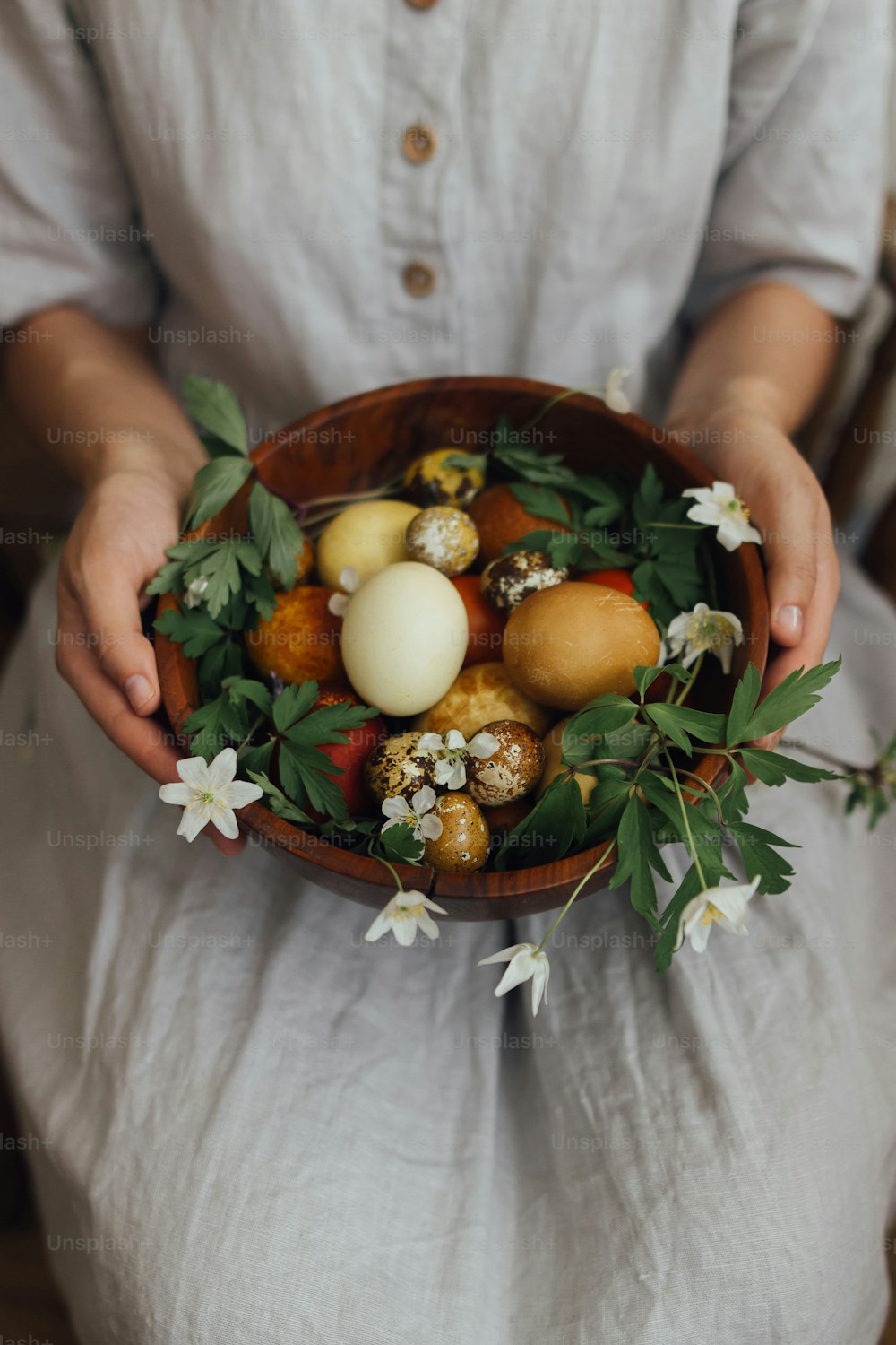 Woman in linen dress holding easter eggs and spring flowers in wooden bowl. Stylish easter and quail eggs in natural dye and spring blooms. Aesthetic holiday. Happy Easter!