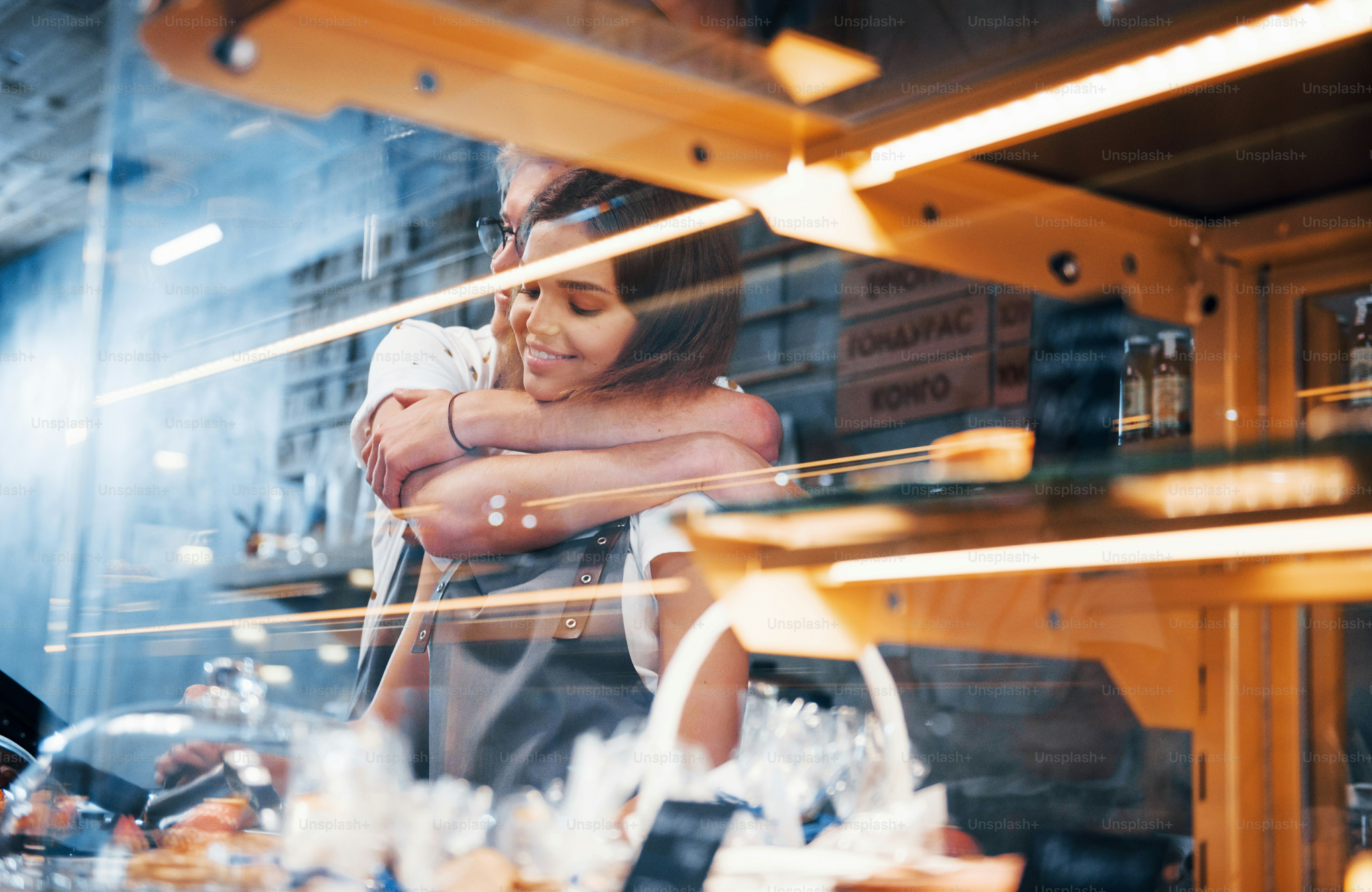 Embracing each other. Behind the glass. Light stripes. Two young cafe workers indoors. Conception of business and service.