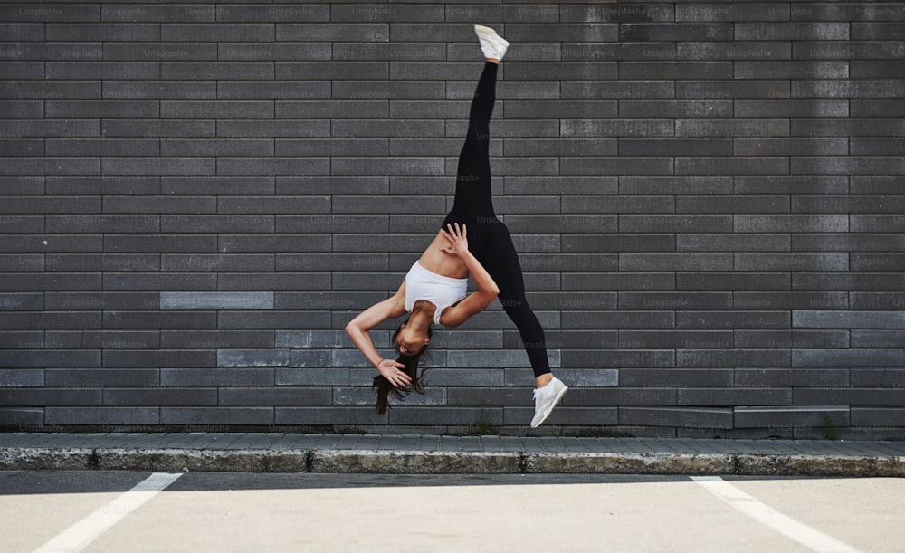 Doing crazy parkour and athletic stunts. Young sportive brunette with slim body shape against brick wall in the city at daytime.