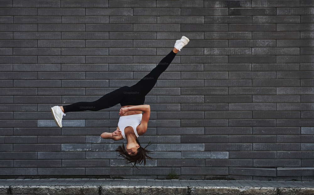 Fare parkour pazzeschi e acrobazie atletiche. Giovane bruna sportiva con la forma del corpo sottile contro il muro di mattoni in città durante il giorno.