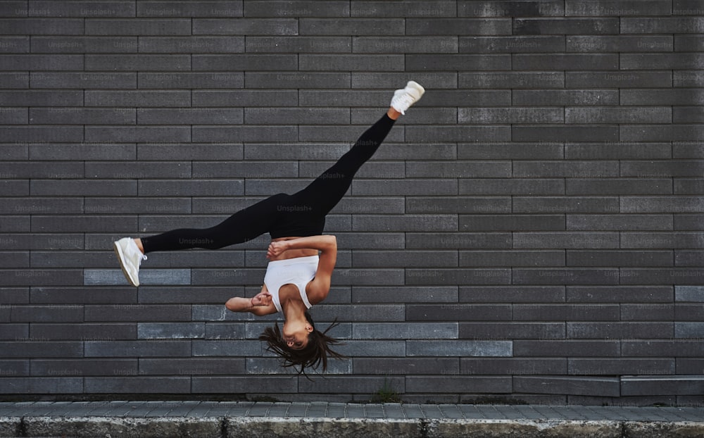 Doing crazy parkour and athletic stunts. Young sportive brunette with slim body shape against brick wall in the city at daytime.