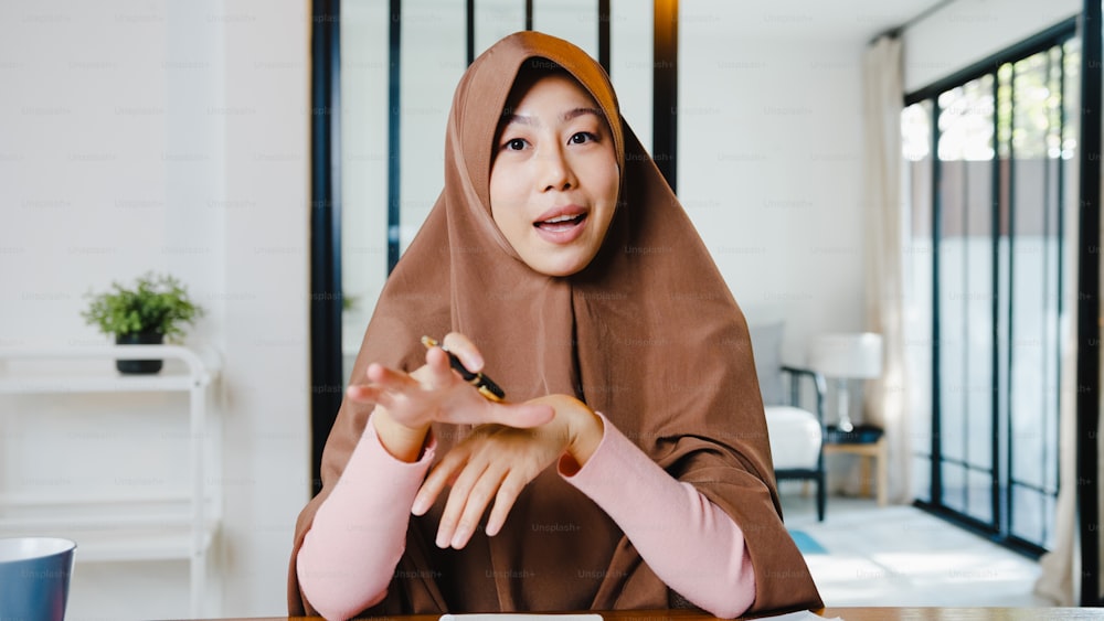 La mujer musulmana de Asia usa el hiyab usando la computadora portátil y habla con colegas sobre el plan en una reunión de videollamada mientras trabaja de forma remota desde casa en la sala de estar. Distanciamiento social, cuarentena por coronavirus.