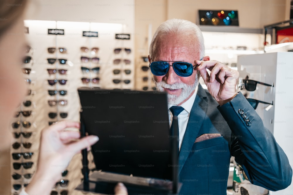 Elegant senior businessman choosing and buying sunglasses in optical store and young female seller helping him to make right decision.