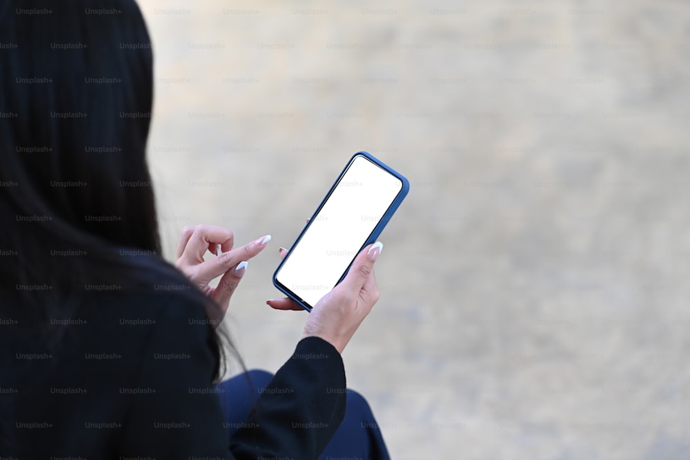 Vista trasera de una mujer de negocios sentada al aire libre y sosteniendo un teléfono inteligente con pantalla blanca.