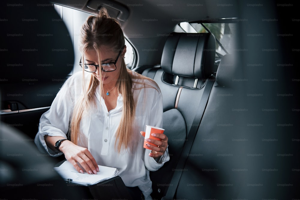 Morning drink in the hand. Smart businesswoman sits at backseat of the luxury car with black interior.