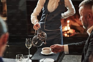 Warm atmosphere. Female waiter pouring hot tea in the white cup. Friends sits in the restaurant with beautiful fireplace in it.