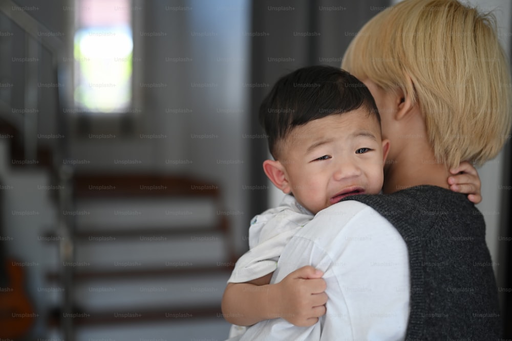 Joven madre asiática sosteniendo a un bebé que llora calmando en un hogar cómodo.
