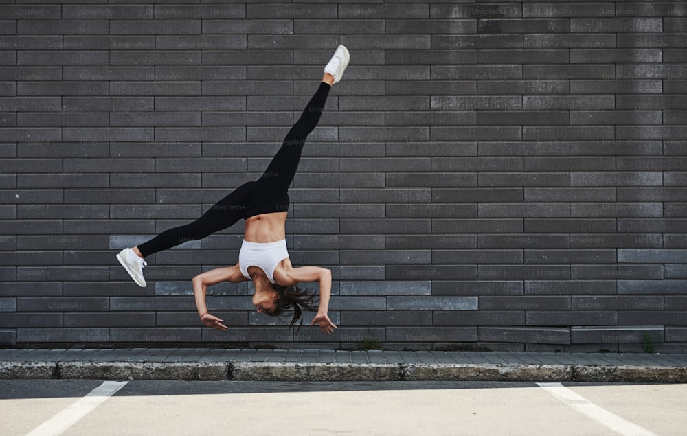 Fare parkour pazzeschi e acrobazie atletiche. Giovane bruna sportiva con la forma del corpo sottile contro il muro di mattoni in città durante il giorno.