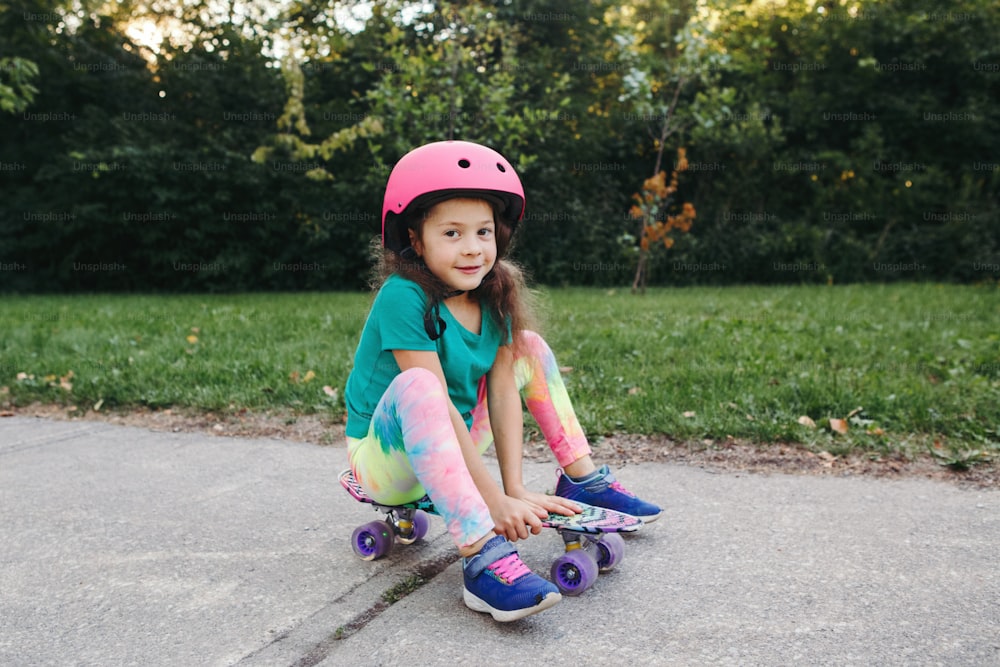 여름날 공원의 도로에서 스케이트보드에 앉아 분홍색 헬멧을 쓴 행복한 미소 짓는 백인 소녀. 계절에 따라 야외 어린이 활동 재미있는 스포츠. 건강한 어린 시절의 생활 방식.