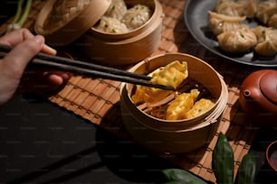 Plan recadré d’une main féminine avec une baguette cueillant des boulettes de Dimsum dans un cuiseur vapeur en bambou sur la table à manger