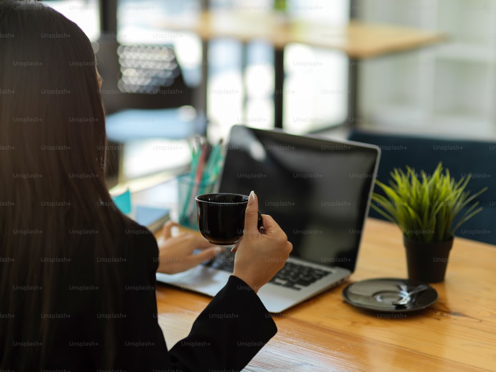 Plan recadré d’une femme d’affaires tenant une tasse de café et travaillant avec un ordinateur portable sur une table en bois dans un café