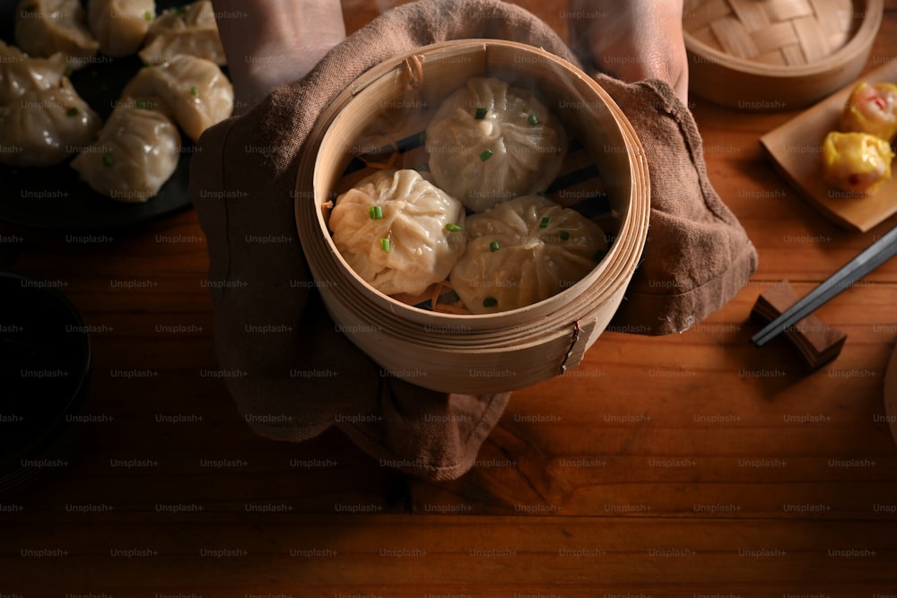 Vue de dessus des mains tenant un cuiseur vapeur en bambou avec des boulettes à montrer à la caméra au-dessus de la table de la cuisine