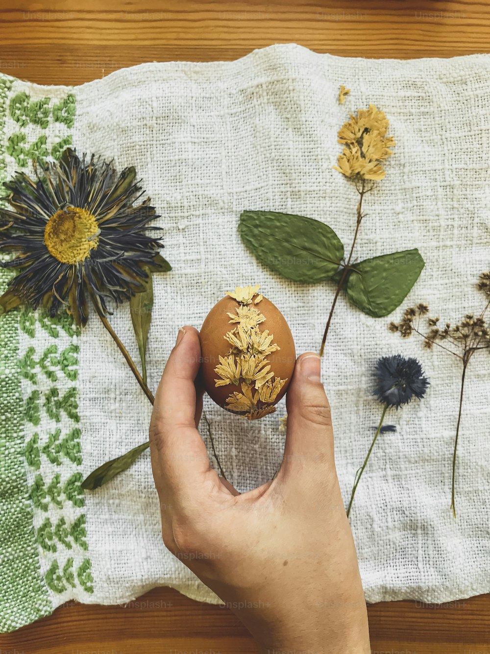 Hand holding Easter egg decorated with dry flower on background of linen napkin and wildflowers. Top view. Creative natural eco friendly decor of easter eggs