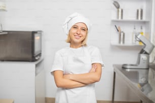 Colpo orizzontale di giovane donna attraente sorridente, proprietario di pasticceria, in posa per la macchina fotografica nella sua uniforme bianca, cappello e grembiule, in piedi nella cucina moderna.