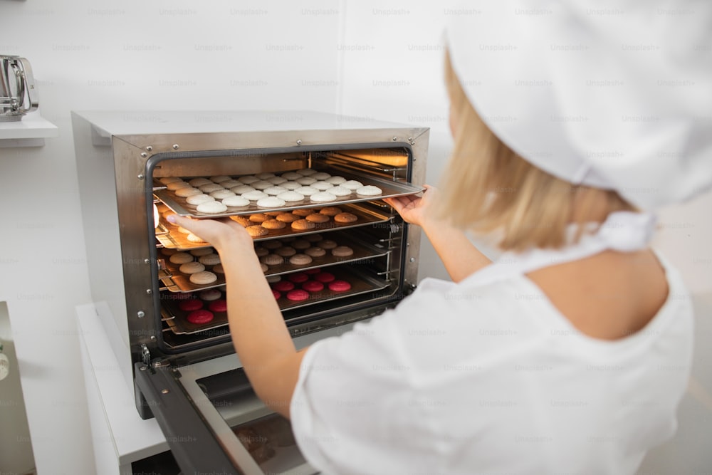 Work of the confectioner. Macaroon baking. Close up back view of woman confectioner putting baking tray with macarons cookies into the oven. Baking cookies and desserts in pastry shop