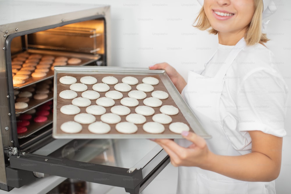 Nahaufnahme einer jungen, fröhlichen Konditorin in der Konditorei, die das Backblech mit weißen Macarons-Keksen hält, bevor sie im Ofen backt. Makronen-Backkonzept.