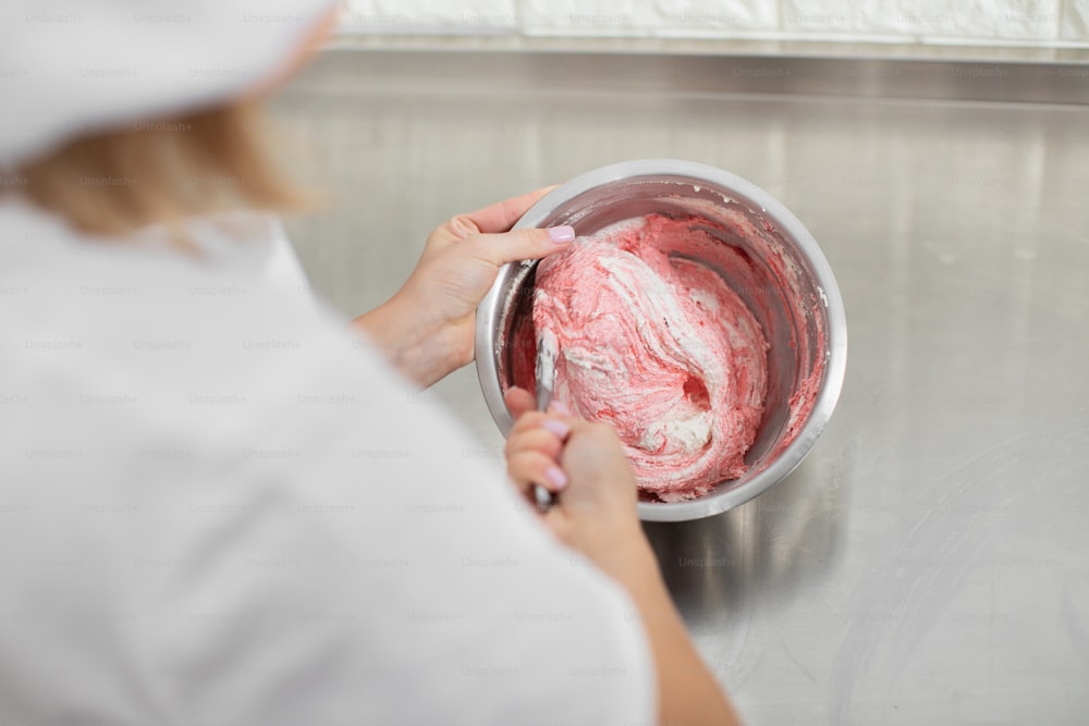 Close up top view of hands of female confectioner in white hat and uniform, mixing red cream in the bowl with spatula. Cream for cake or macarons. Cooking, desserts and pastry shop concept.