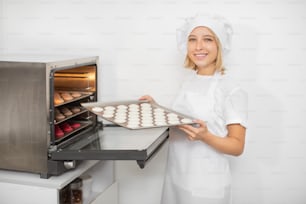 Confectionery shop, macarons baking. Pretty woman confectioner in white uniform and hat, holds a batch of macarons on baking tray, ready to put it into the oven