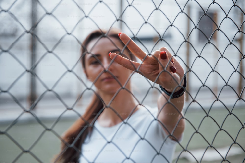 Brunette stands behind the fence and shows gesture using two fingers.