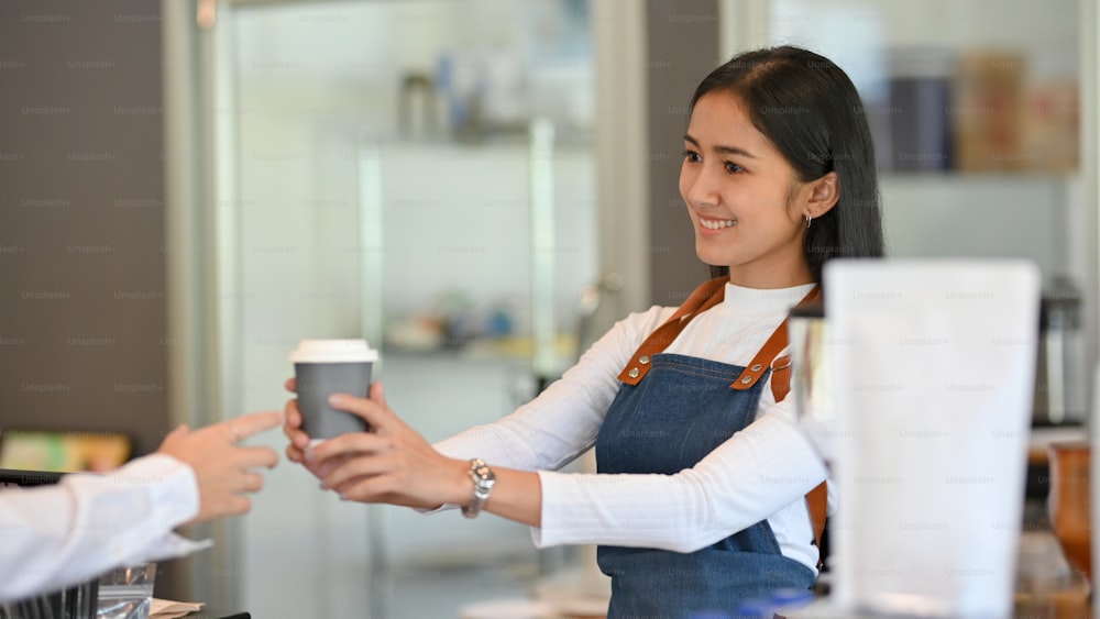 Young beautiful barista handing the coffee cup over to her customer in coffee shop