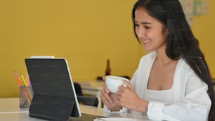 Cropped shot of young beautiful female holding coffee cup and smiling while looking on digital tablet
