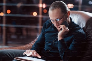 Credit card in the hand and tablet on the legs. Stylish businessman in eyewear works alone in the office at nightime.