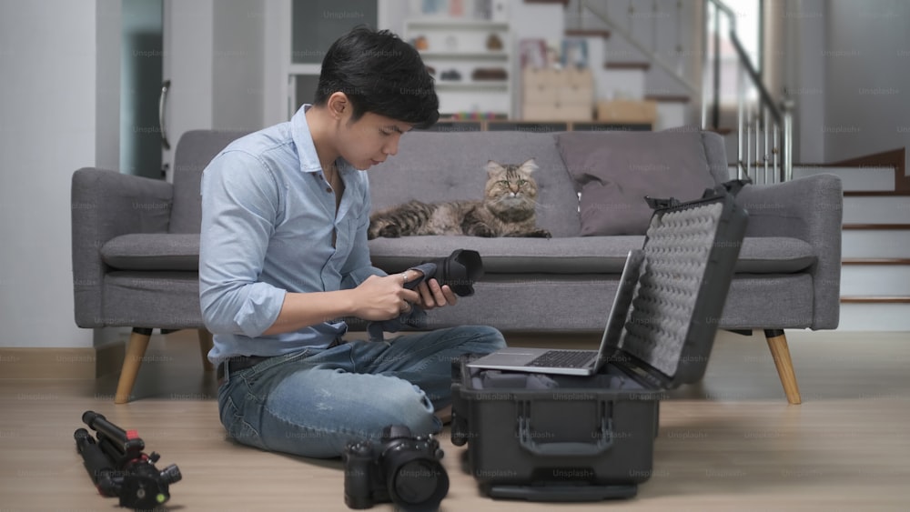 Side view of young man photographer put camera accessories in the bag while sitting on wooden floor at home.
