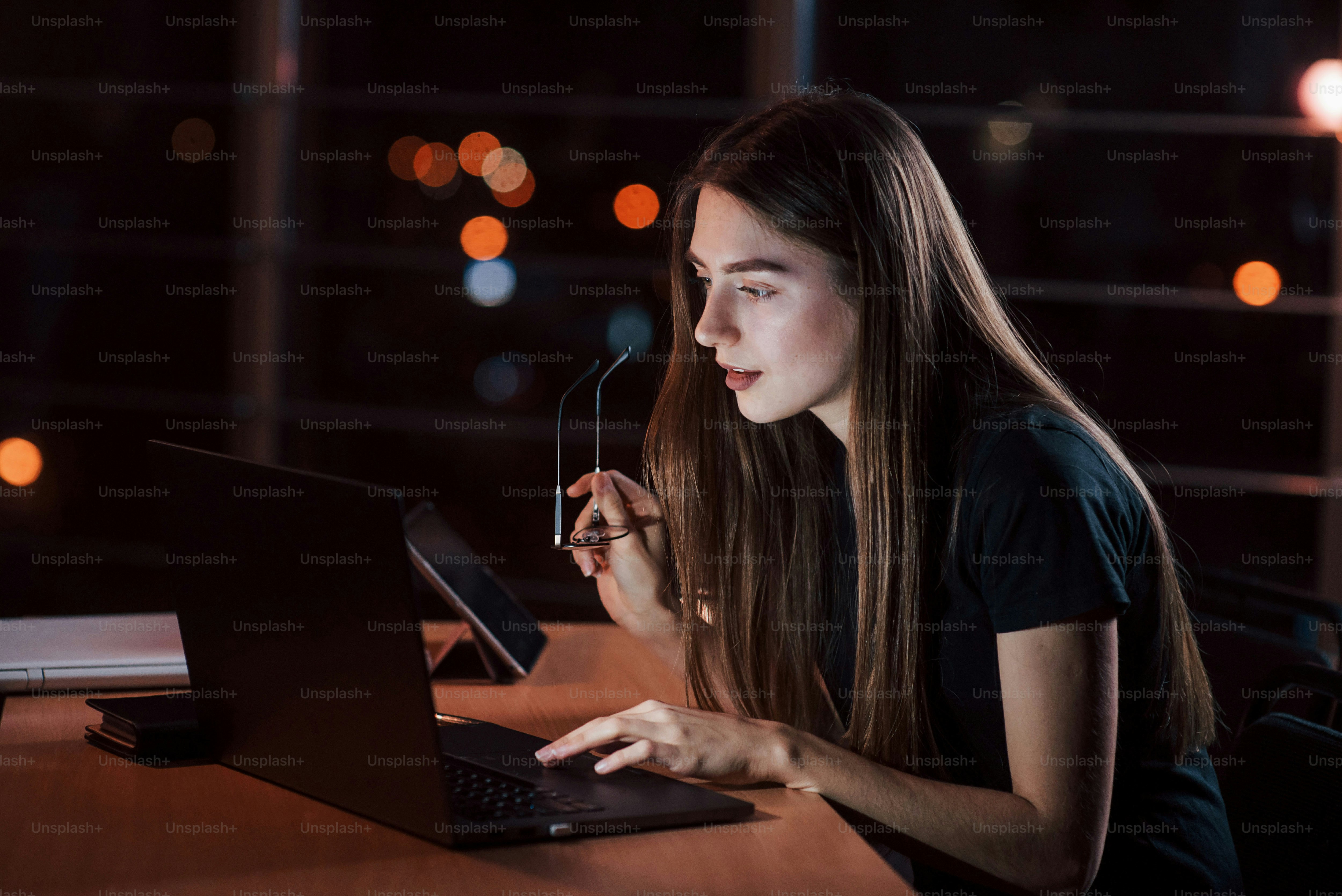 Holds eyewear in the hand. Beautiful blonde businesswoman works alone in the office at nightime.