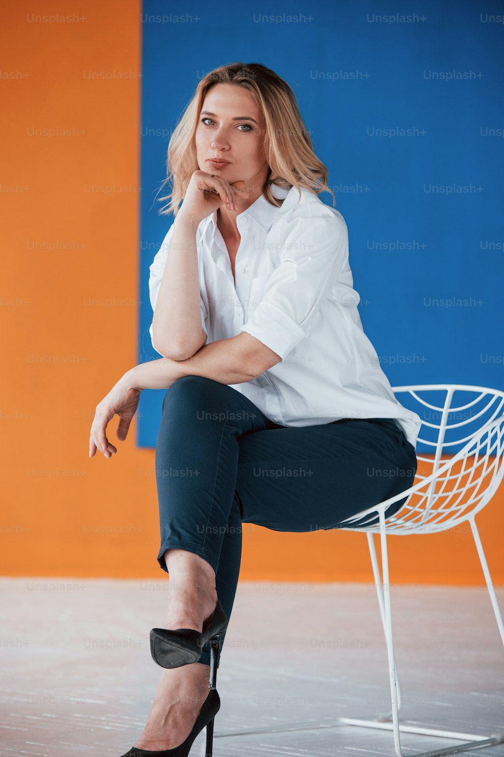 Sitting on white chair. Businesswoman with curly blonde hair indoors in room with orange and blue colored wall.
