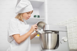 Jovem loira, confeiteira profissional, vestindo avental branco e chapéu de chef, cozinhando no interior aconchegante da cozinha leve, usando misturador ou processador de alimentos para chicotear claras de ovos.