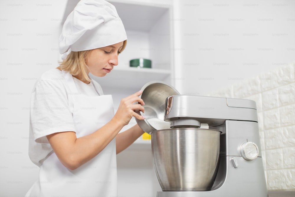 Young blond girl, professional confectioner, wearing white apron and chef's hat, cooking in cozy light kitchen interior, using mixer or food processor for whipping egg whites.