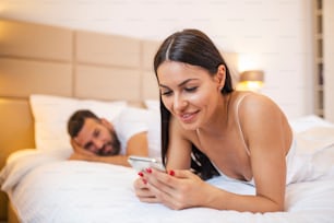 Addicted young woman on bed texting while man looks angry. Young couple having a problem. Woman is chatting using a smartphone while her boyfriend is bored in bed