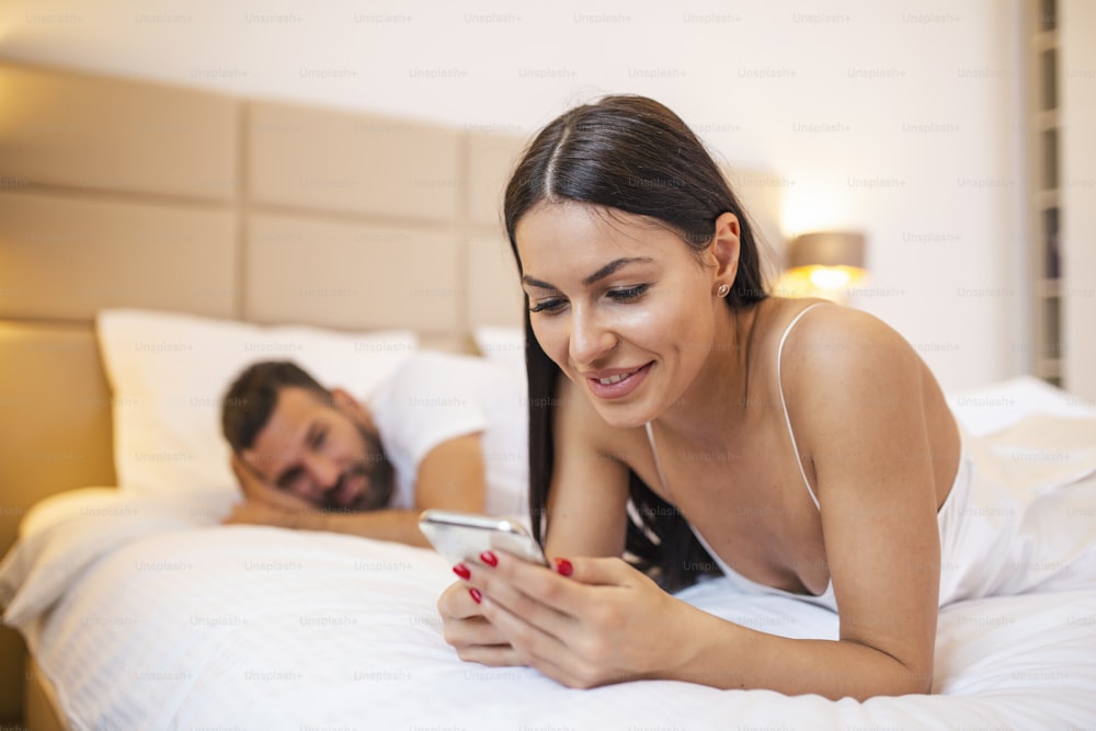 Jeune femme accro au lit en train d’envoyer des SMS pendant que l’homme a l’air en colère. Jeune couple ayant un problème. Une femme discute à l’aide d’un smartphone pendant que son petit ami s’ennuie au lit