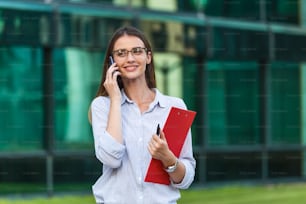 Young businesswoman speaking by smartphone while standing by minimal office building interior, copy space. Portrait of an attractive businesswoman standing in front of windows in an office building