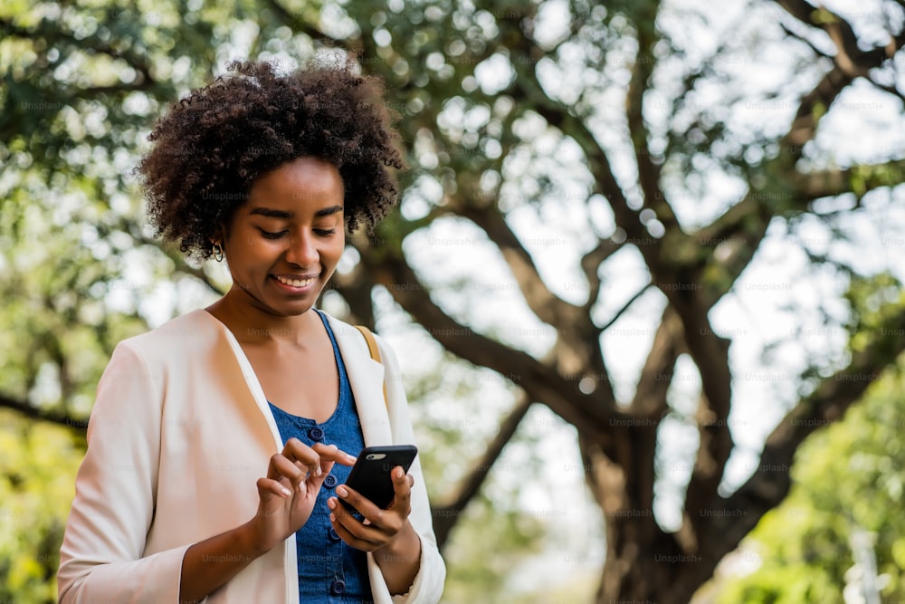 Portrait of afro business woman using her mobile phone while standing outdoors at the park. Business and urban concept.
