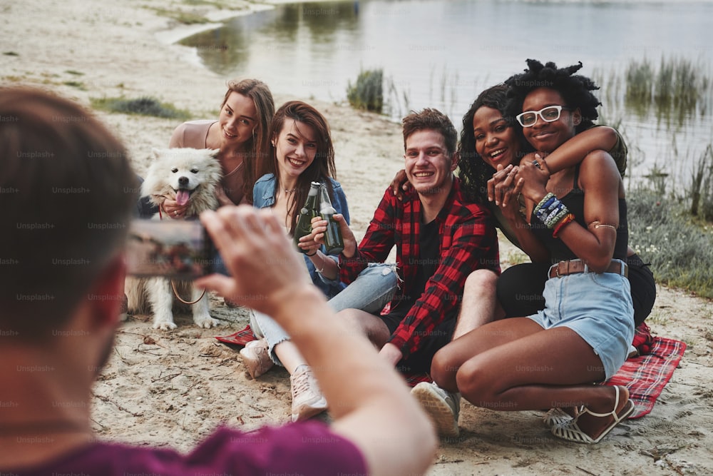 Everyone looks in the camera. Group of people have picnic on the beach. Friends have fun at weekend time.