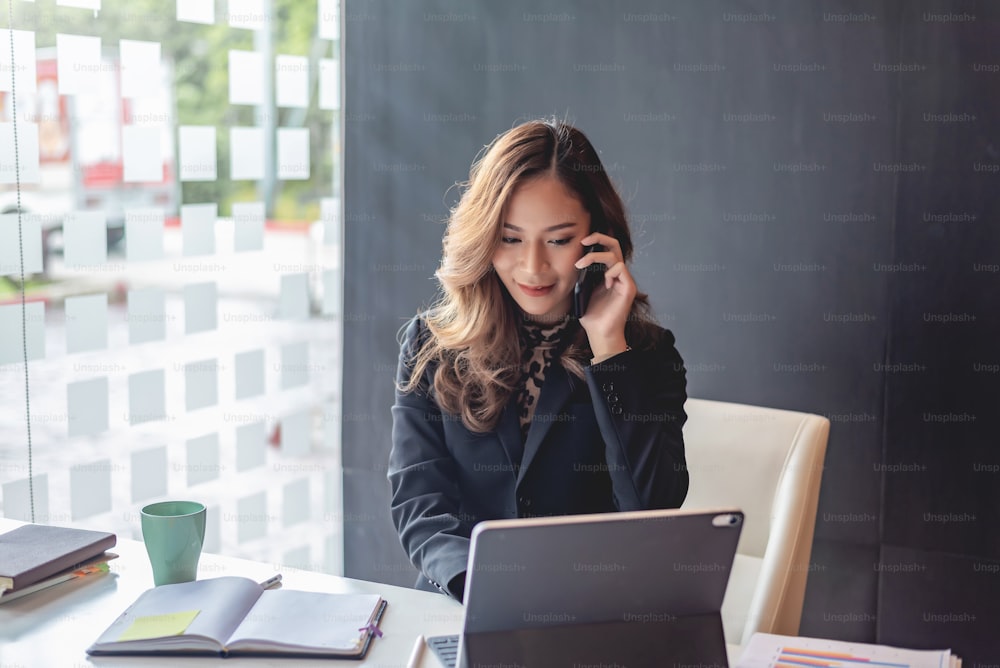 Attraente giovane donna asiatica che parla al telefono cellulare e in piedi al suo posto di lavoro in ufficio.