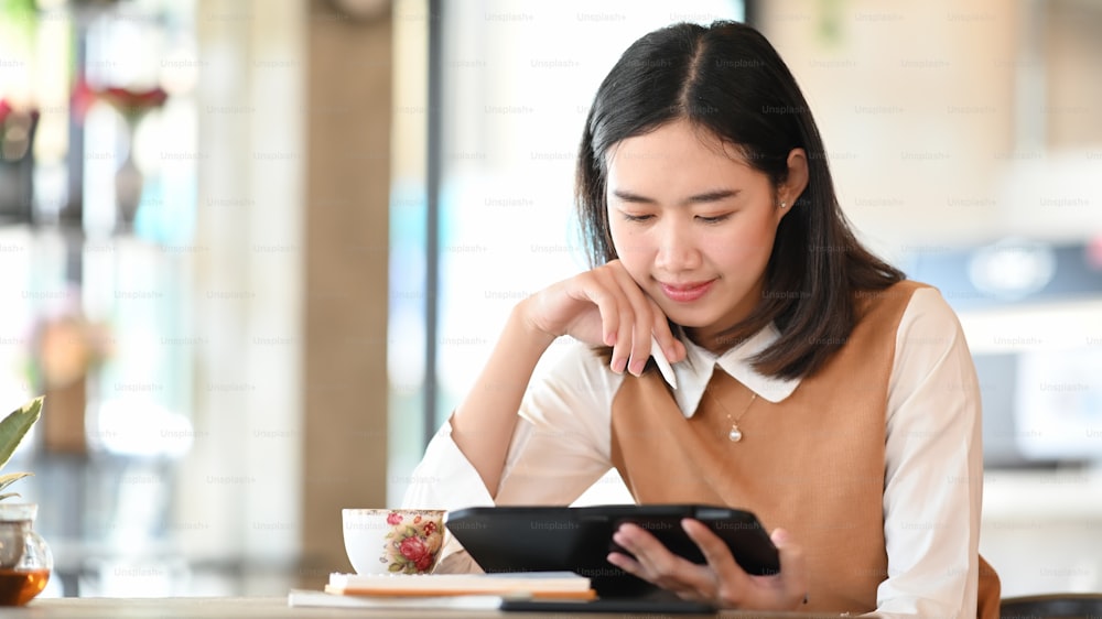 Junge Frau sitzt im Café und liest E-Mails auf digitalem Tablet.