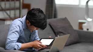 Joven hombre casual que usa una tableta digital y trabaja en una computadora portátil mientras está sentado en el sofá.