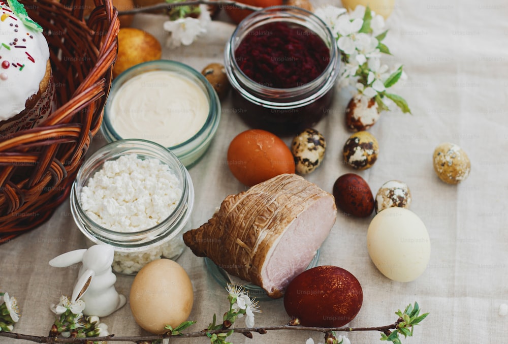 Delicious Easter food, stylish easter eggs, beets, cheese, butter, ham, homemade Easter bread and wicker basket with blooming spring flowers on rustic table, orthodox traditions. Happy Easter!