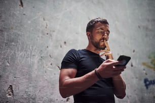 Messaging by using phone. Portrait of man in black shirt smoking on the background of old cracked wall.