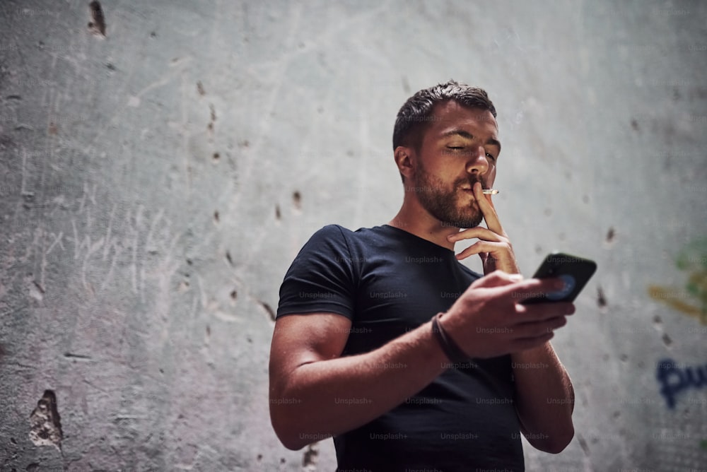 Messaging by using phone. Portrait of man in black shirt smoking on the background of old cracked wall.