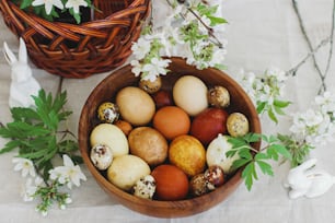 Happy Easter! Stylish easter eggs in wooden bowl on rustic table with bunny figurines, wicker basket and spring blooming flowers. Natural dyed eggs in yellow and red colors