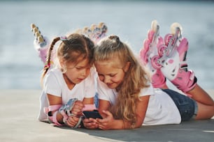 Using smartphone. Two little girls with roller skates outdoors near the lake at background.